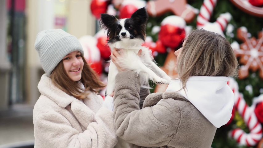 happy beautiful mom and daughter in a fur coat with dog Papillon near Christmas tree on the street. Walks in winter with pets. decorating parks for the new year. posing for a winter family photo shoot