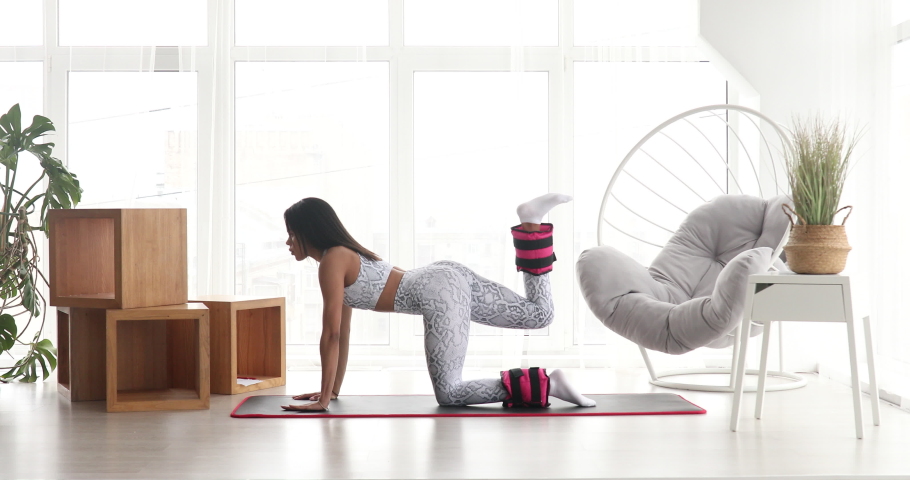 Pretty fitness black woman in sportswear practicing donkey kick exercise with ankle weight for glutes strength training at home during covid-19. | Shutterstock HD Video #1064928898