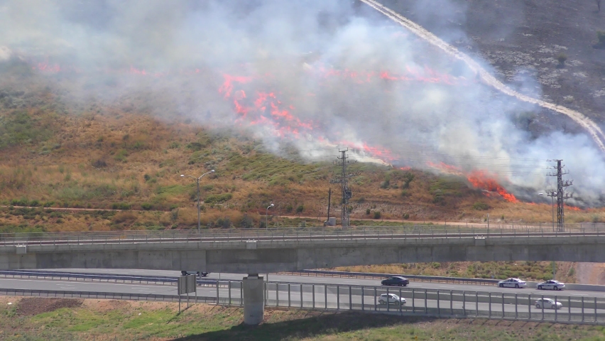 Mount carmel is burning and there is a lot of smoke from the strong wind, cars driving below in nearby highway
