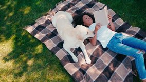 African american girl reading book while lying on plaid blanket near labrador