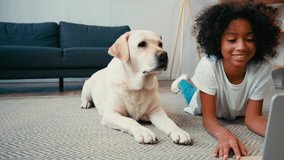 African american girl lying on carpet near laptop and dog wagging tail at home