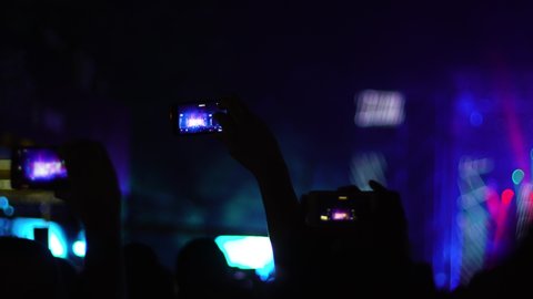 Abstract silhouette people in a crowd, hands up, dancing and holding smart phones. Out of focus stage. Handheld video