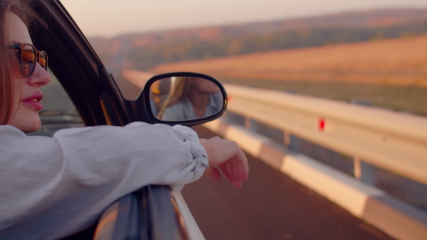 Modern wanderlust travel by car. The side rearview mirror is a reflection of a young woman sitting in an antique cabriolet and looking at the sunset. Her hand is on the front door of the car.
