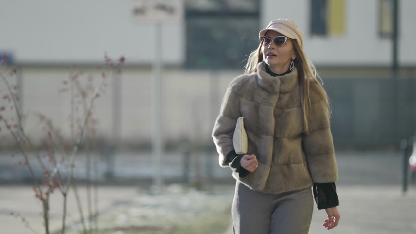 rich young woman dressed luxury expensive fur jacket is walking on street at sunny weather