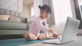 Little asian girl doing yoga plank and watching online lessons on laptop, exercising in the kitchen. A child plays sports during the COVID-19 pandemic.
