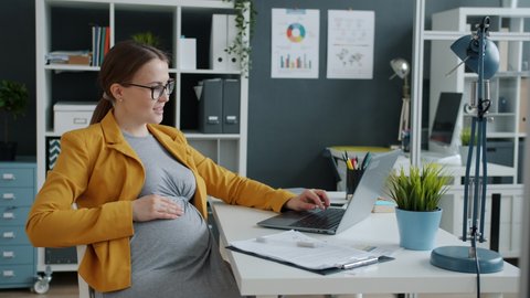 Beautiful woman expecting baby working with laptop computer in modern office typing and storking belly sitting at desk alone. Pregnancy and workplace concept.