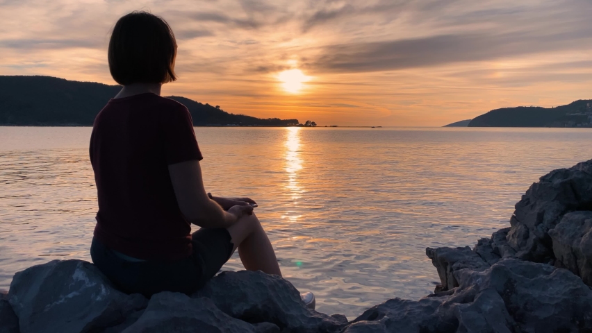 Young romantic woman sits on rock on seashore and admires spectacular seascape and fiery sunset. She takes out her mobile phone to take photos of beautiful view