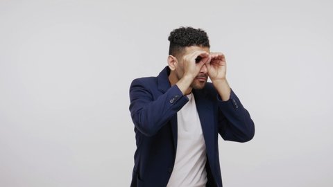 Funny bearded man in suit spying holding fists near eyes and looking through holes, pretending to watch in binoculars, zooming to see better, having fun. Indoor studio shot isolated on gray background
