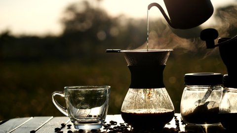 Drip coffee brewing while outdoor camping,pouring hot water from kettle over coffee ground extracting coffee through filter with nature steam smoke of coffee in beautiful sunrise background in morning