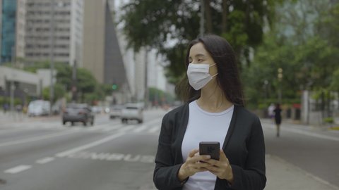 Front view of  woman in the city streets during the day, wearing a face mask against air pollution and coronavirus Covid19, standing and using a smartphone
