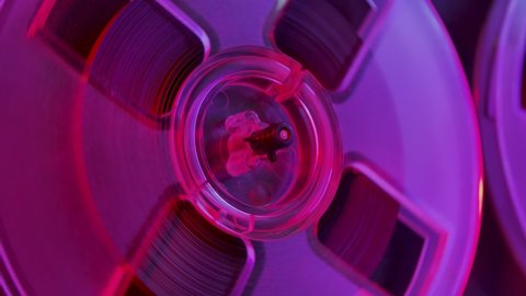 Detailed macro shot of a transparent reel rotating on a vintage analog reel to reel tape recorder. Bobbin with retro tape in the light of the club violet-pink lights. Retro party. Close up. Slow