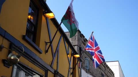 British Union Jack and Welsh flags flying on yellow Tudor style house wall against blue sky.