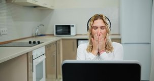 Bored corporate blonde worker in white blouse covers mouth with hands and yawns sitting at online conference in home kitchen