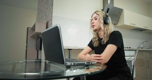 Serious blonde student in black t-shirt and headphones talks looking into laptop at glass table during covid isolation at home