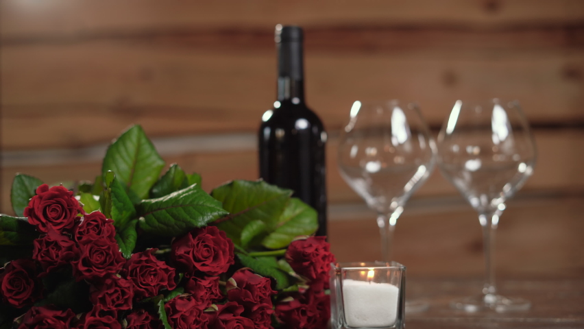 a bottle of wine and two glasses are on the table, a candle is burning nearby. A bouquet of red roses, a woman