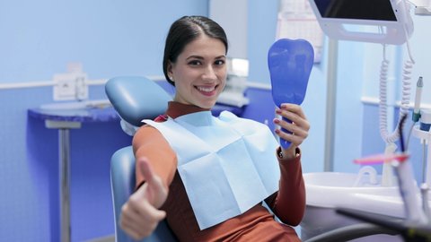 Female patient in the dental clinic is satisfied with the results of the procedures, looking at mirror and smiling. Visiting dentist. Healthcare and medicine concept