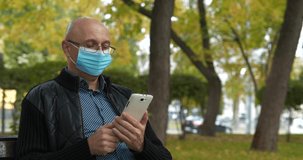 Senior man in medical protective mask sits on bench and uses his smartphone. Covid concept health and safety, coronavirus quarantine, next wave covid virus protection. Cinema 4K 60fps video