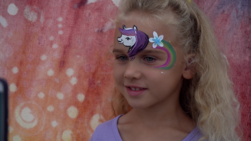 Excited little girl enjoy painting on her face. Pony rainbow and flower painted on child forehead. Preschooler girl looking at phone with her photo. Camera motion shot with gimbal.