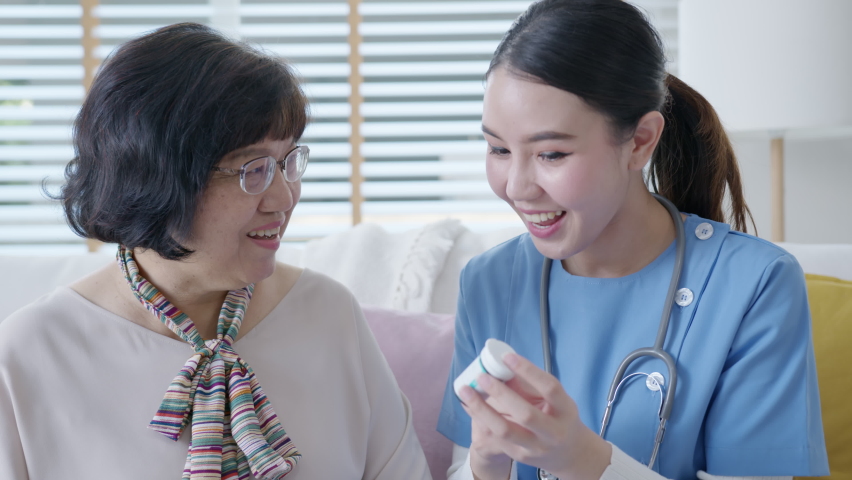 Young caregiver in scrubs uniform showing medicine bottle to old elderly asian woman in home visit care nursing service.  Asian senior with assisted living medication monitoring sit and talk at home. | Shutterstock HD Video #1064997103