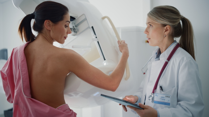 Friendly Female Doctor with Tablet Computer Explains Mammogram Procedure to a Topless Adult Female Patient Undergoing Mammography Scan. Healthy Woman Does Cancer Prevention Routine in Hospital Room. Royalty-Free Stock Footage #1064997862