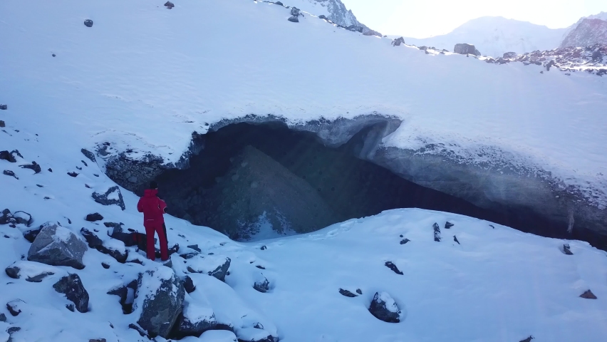 A climber looks at the entrance to an ice cave. Frozen snow mountains and glacier. Entrance looks like a dolphin. High peaks, clear blue sky and big rocks. An extreme journey. Lots of snow underfoot
