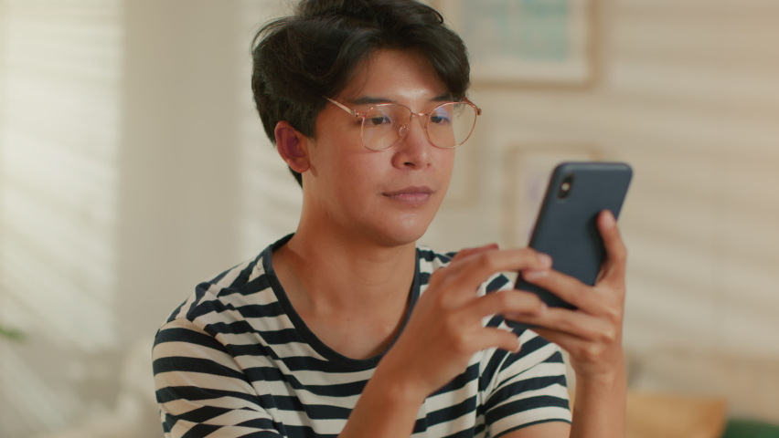 Excited Asian Man having good news on phone smile and enjoy success on phone. Joyful Man reading good news making yes gesture celebrating good news while sitting in living room at home | Shutterstock HD Video #1065003838