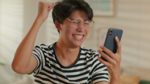 Excited Asian Man having good news on phone smile and enjoy success on phone. Joyful Man reading good news making yes gesture celebrating good news while sitting in living room at home