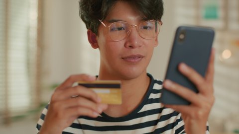 Attractive Young Asian Man is making online payment by using a credit card, hand typing number of Card on smartphone device sitting in living room at home. Buying online, Internet shopping Concept
