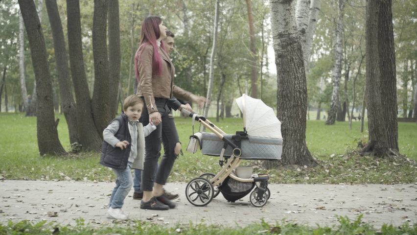 Side view of relaxed young family strolling in spring or summer park and talking. Wide shot of Caucasian man with baby stroller and woman holding hand of little boy walking outdoors.
