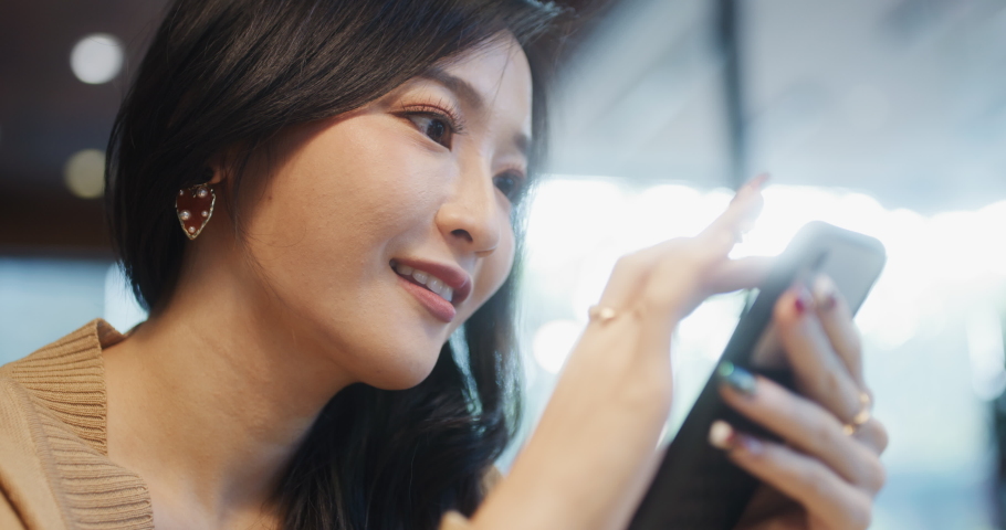Beautiful young Asian woman using smartphone in coffee shop, happy smiling. People lifestyle, internet communication technology, or online shopping concept. Handheld slow motion. Shot on RED Komodo 6K Royalty-Free Stock Footage #1065017428