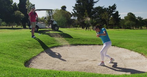 Two caucasian women playing golf wearing face masks one taking shot from bunker. golf cart and clubs in background. golf sports hobby healthy lifestyle hygiene during covid coronavirus pandemic.