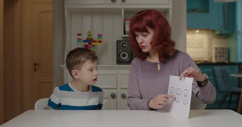 Autism psychologist teaching kid with autism emotions. School boy with autism learning with mother at home. Learning happy, surprise and upset feelings by card. 
