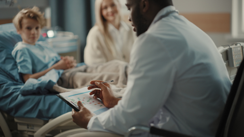 Hospital Ward: Handsome Young Boy Resting in Bed with Caring Mother Visits to Support Him, Friendly Doctor Talks, Gives Advice. Happy Smiling Patient Recovering after Sickness or Successful Surgery