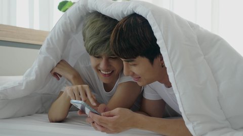 Lovely Asian gay couple laying on bed at day time, cover their heads with white blanket, talking, using smartphone, smiling, and flirting. Indoors, handheld, LGBT and same sex relationship concept