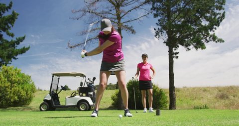 Two caucasian women playing golf one taking shot from bunker. golf cart and clubs in background. golf sports hobby healthy lifestyle.