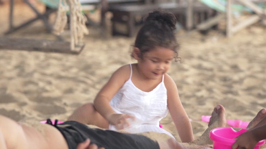 Happy Asian family holiday vacation. Adorable little daughter sitting on sand beach and playing beach toy and sand with her father and sister. Smiling cute child girl having fun on the beach in summer