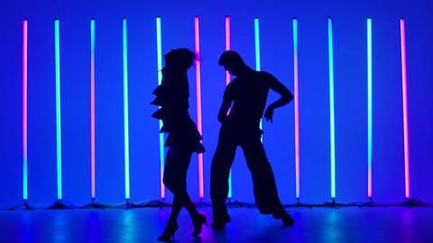Professional dancers dance rumba in the studio against the backdrop of bright neon tubes. A guy and a girl practice the steps of the dance. Dark silhouettes of dancers. Slow motion.