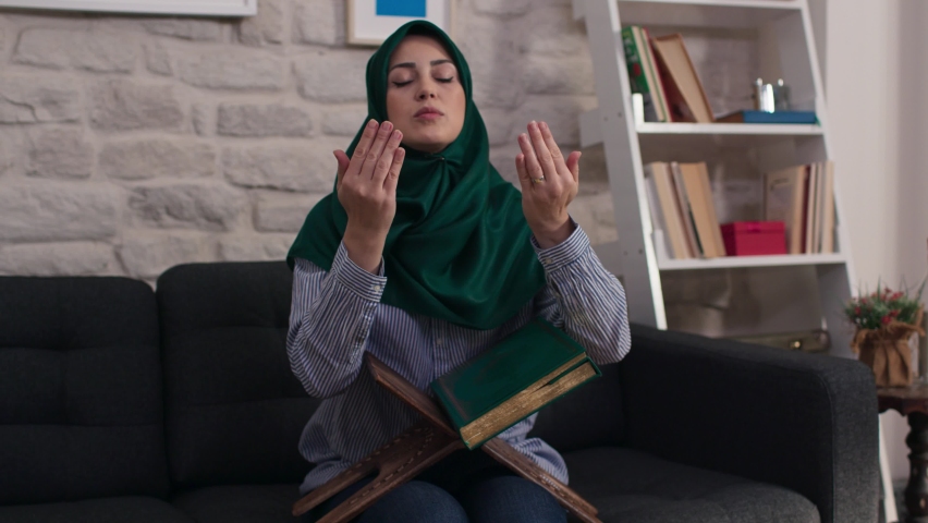 The young Muslim woman reading the Koran at home raises her hand and prays. Praying Muslim woman.