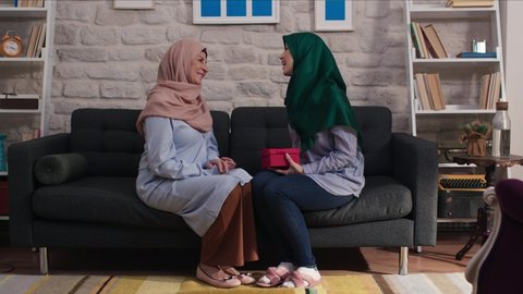 Loving adult daughter in hijab is giving a gift box to her mother. The young woman gives a gift to her mother's day, hugs and congratulates her mother at home.