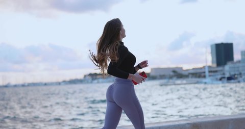 Attractive sexy young sportswoman enjoying evening running workout along the beach listening music in earphones holding smartphone.