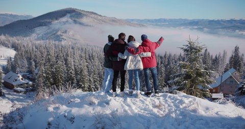 Caucasian happy young people gathering on mountain top hugging each other celebrating winter holiday on sunny weather. Winter resort. Vacation concept.