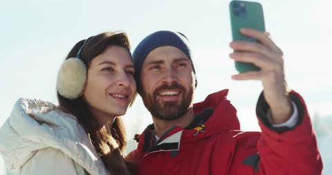 Amazing caucasian young couple holding smartphone taking pictures on camera enjoying winter sunny weather holiday ski resort outdoors. Family selfie.