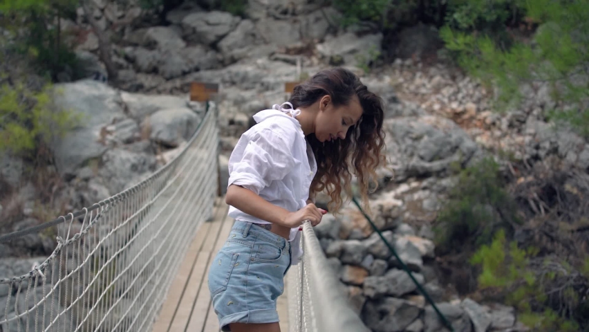 Brunette with long loose flowing curly hair stands on suspension bridge and looks downward holding hands on grey rope handrails against large rocky hill slow motion.