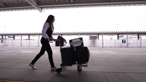 Woman push baggage dolly full of suitcases, walk near airport terminal. European lady arrive to or depart from domestic terminal of Japanese airport. Tracking shot, side view