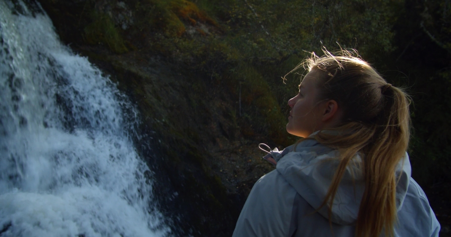 Slow motion. Young attractive Finnish woman in a grey coat looking up in awe at a running waterfall in the Scottish Highlands while travelling the North Coast 500. Scotland, Tourism, Adventure.