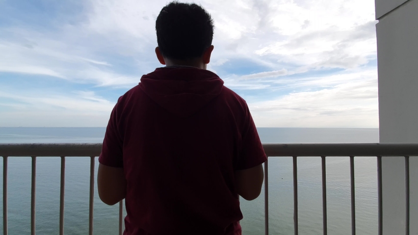 Silhouette of a boy enjoys morning sunrise or sunset from his hotel balcony with calm blue ocean at the background, serene scene