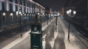Special equipment removes snow on a beautiful old city street. Night, snowfall, the street is decorated with New Year's garlands. In the foreground is a clock with a sculpture. Video from a drone.