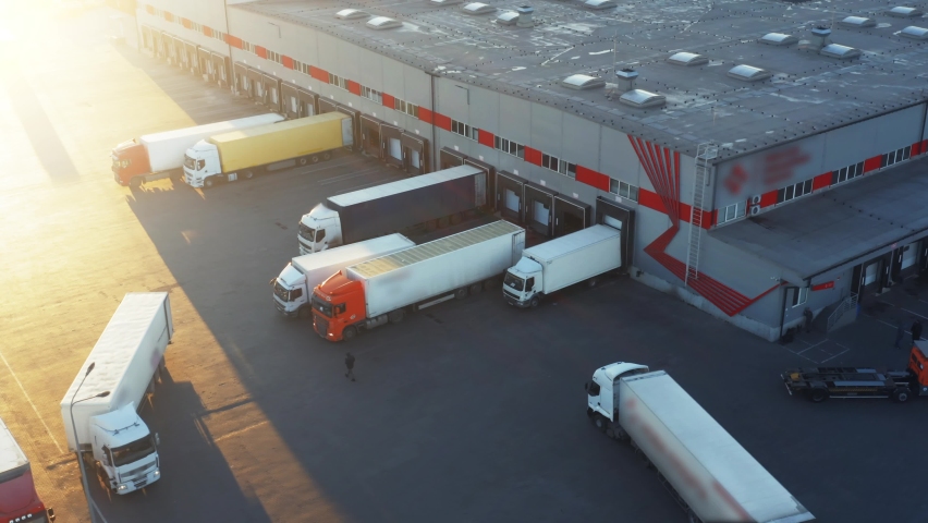 Semi-trailer trucks stand at the ramps of warehouse in the logistics park with loading hub and wait for load and unload goods at sunset. Aerial view Royalty-Free Stock Footage #1065049711