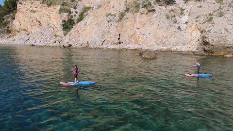 Young couple is engaged in active recreation during vacation at seaside resort. Man and woman row oars standing on SUP boards and swim along cliffs, shooting with drone from height