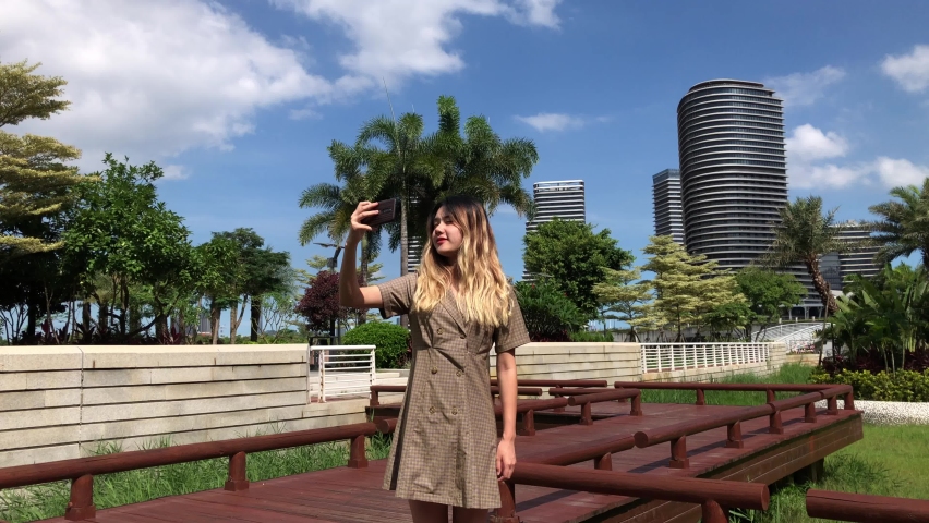 Chinese girl standing outside in park taking selfies. China woman outdoors shooting self shot with pro camera, posing and mugging. Cute girl takes photos with point and shoot vlog camera.